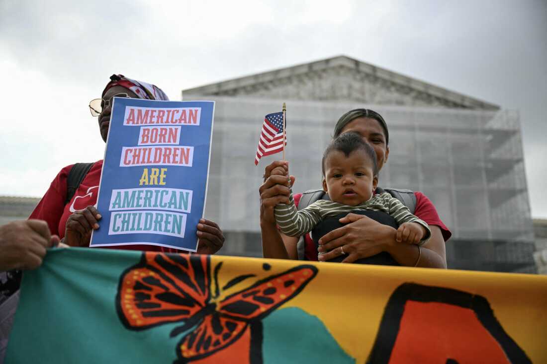 Olga Urbina holds her 9-month-old son, Ares Webster, in a front-pack baby carrier at a protest in front of the Supreme Court building. Ares holds a small American flag. Another protester holds a sign that says, "American Born Children Are American Children."