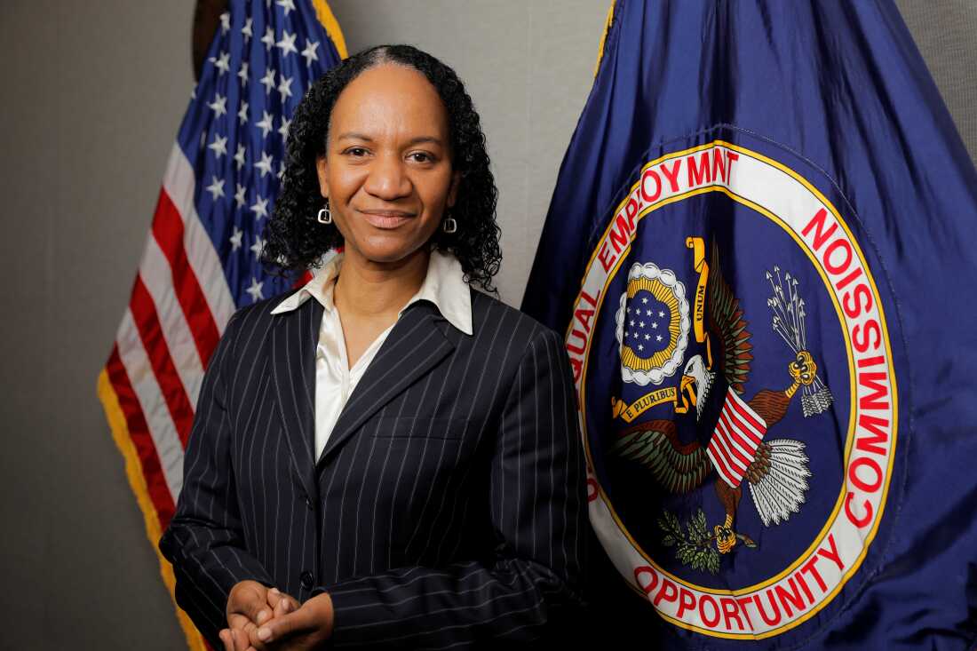 Charlotte Burrows, then-EEOC chair, in 2021. She is a Black woman standing in front of an American flag and the EEOC flag, which is blue and has an eagle on it.