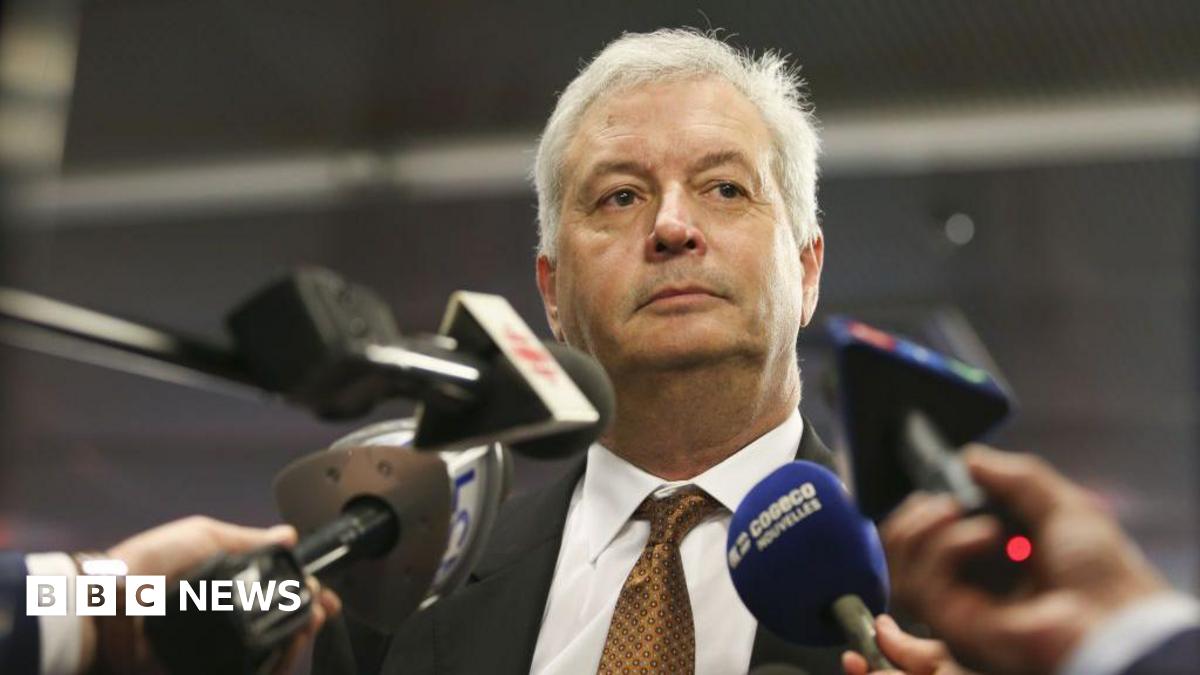 An image showing Michael Rousseau surrounded by media microphones at a scrum in Montreal, pictured in 2021. He has short white hair, and is wearing a suit and copper patterned tie.