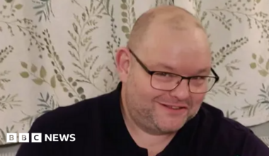 A man smiles into the camera. He is wearing black glasses and a black top. Behind him are white curtains with details of green leaves and branches.