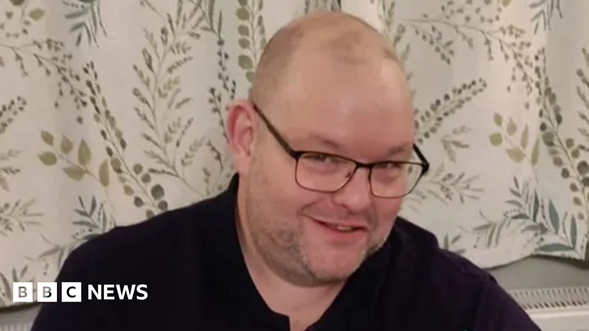 A man smiles into the camera. He is wearing black glasses and a black top. Behind him are white curtains with details of green leaves and branches.