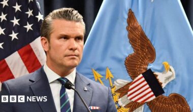 Hegseth in navy suit with red and blue tie at a podium with Pentagon seal behind him alon with flag and people raising hands in front of him