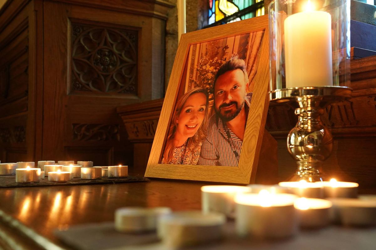 An indoor setting features a framed portrait of a couple displayed on a wooden table, surrounded by several lit candles and a candle holder, creating a somber and contemplative atmosphere.