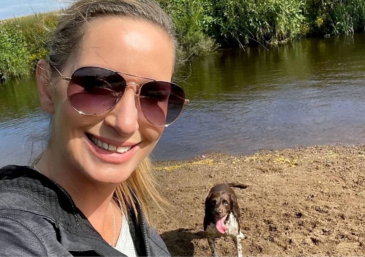 A woman wearing sunglasses and a backpack is smiling near a river with a dog in the background.