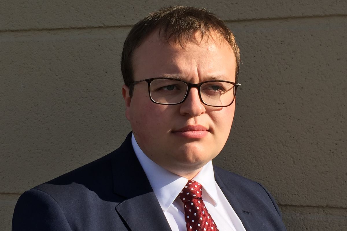 A white man in a suit and tie. He has glasses and short dark hair