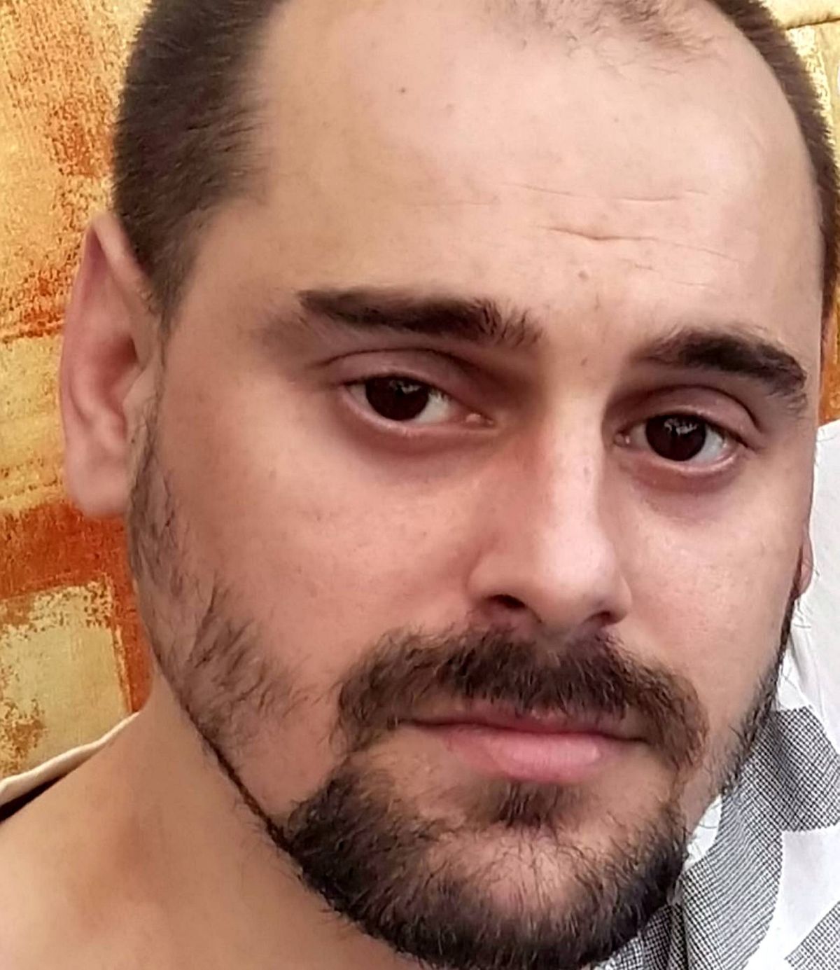 An individual with a beard and mustache is featured in a close-up, with a focus on the facial hair and the upper part of the torso. The person's expression is neutral, and the background is intentionally blurred to emphasize the facial details.