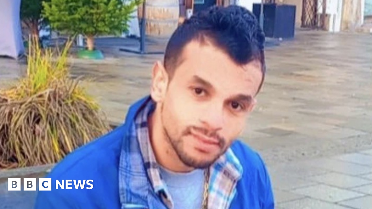 A man in a blue jacket and checked shirt sat down on a bench outside. He is holding a phone with headphones plugged in