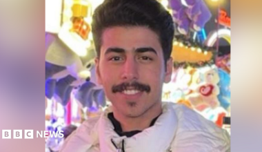 Mohammed Algasim: a dark-haired man with a dark moustache. He is wearing a white anorak-style coat and is standing in front of a multi-coloured background