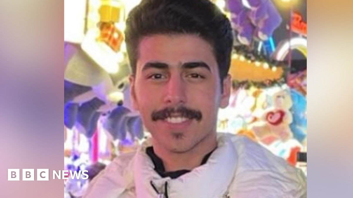 Mohammed Algasim: a dark-haired man with a dark moustache. He is wearing a white anorak-style coat and is standing in front of a multi-coloured background