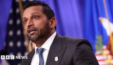 close-up of Kash Patel speaking, wearing a suit and tie in front of American flag