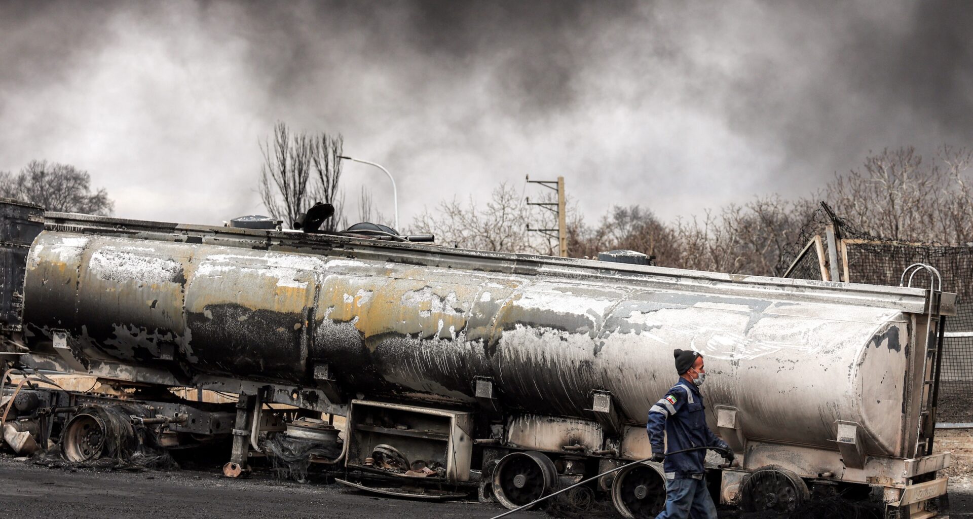 An Iranian civil defence member walks with a hose next to a destroyed fuel tanker vehicle near an ongoing fire following an overnight airstrike on the Shahran oil refinery in northwestern Tehran on March 8, 2026.