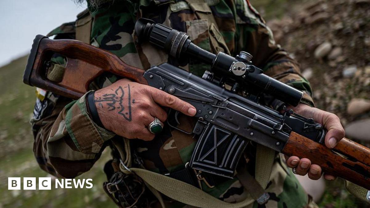 A man in camouflage gear grips an assault rifle on a rocky hillside.