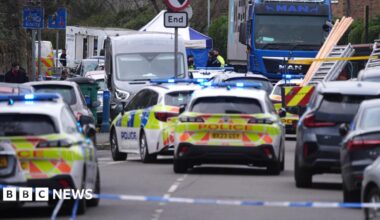 Police cars and other vehicles at the scene on a residential street. A police cordon can be seen at the front.