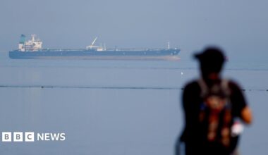 A woman with dark hair and a multiple colour jumper siting on a green sofa inside her living room.