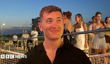 Will Bailey looks to the side as he smiles. He is sitting down wearing a black shirt. People can be seen walking on a walkway behind him.