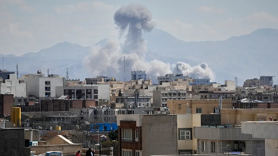 People watching from a rooftop as a large plume of smoke rises over Tehran, Iran.