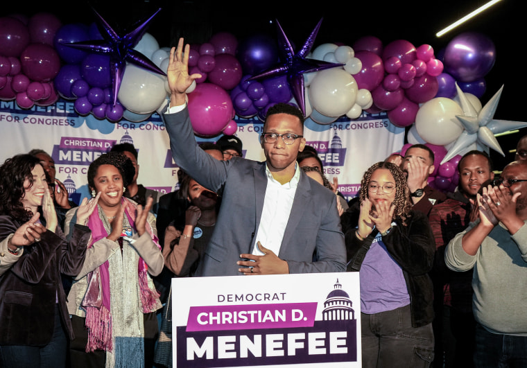 Christian Menefee raises his right hand while standing in front of a campaign sign bearing his name at an election party.