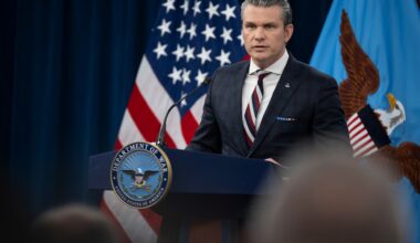 A standing man in a business suit speaks from a lectern, with an American flag and a blue flag with an eagle on it in the background.