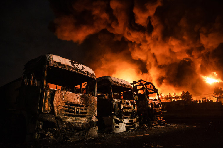 Charred vehicles seen outside near plumes of smoke and fire in the distance