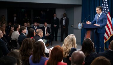 A man in business attire stands behind a lectern and speaks to a seated audience; behind him is an American flag.