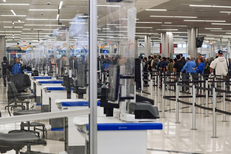 Empty TSA desks behind plastic barriers at an airport. A line of people is visible in the background.