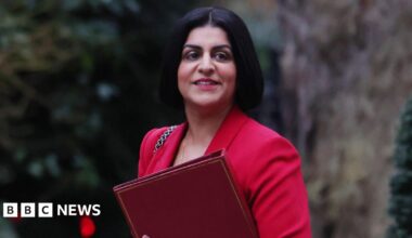 Shabana Mahmood walks through a dormitory room which contains bunk beds to her left and right. She is wearing a black coat and black shoes.