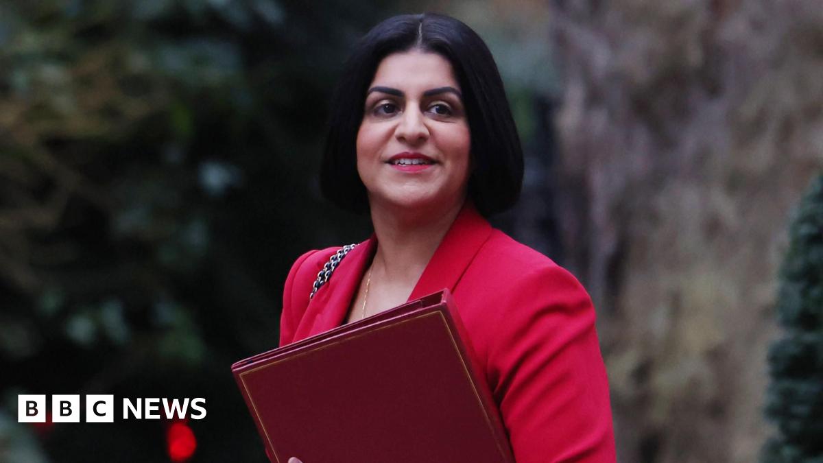 Shabana Mahmood walks through a dormitory room which contains bunk beds to her left and right. She is wearing a black coat and black shoes.