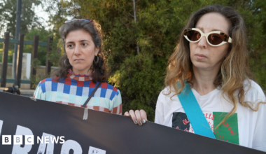 Three people standing behind a black sign they are holding up that reads white and red: "British Bases Out."