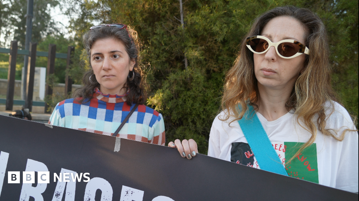 Three people standing behind a black sign they are holding up that reads white and red: "British Bases Out."