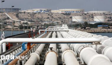View of Kharg Island oil terminal, showing a row of pipes stretching into the distance towards a raised area of ground where several silos sit, one of them has the words "Persian Gulf" on its side.