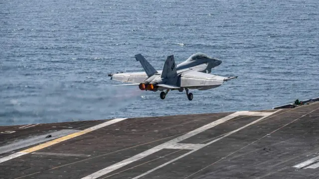 A US strike jet takes off from an aircraft carrier sailing on calm blue water.
