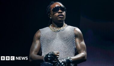 Gunna, dressed in gold and silver chains wears a silver vest top, black gloves and sunglasses while holding a mic
