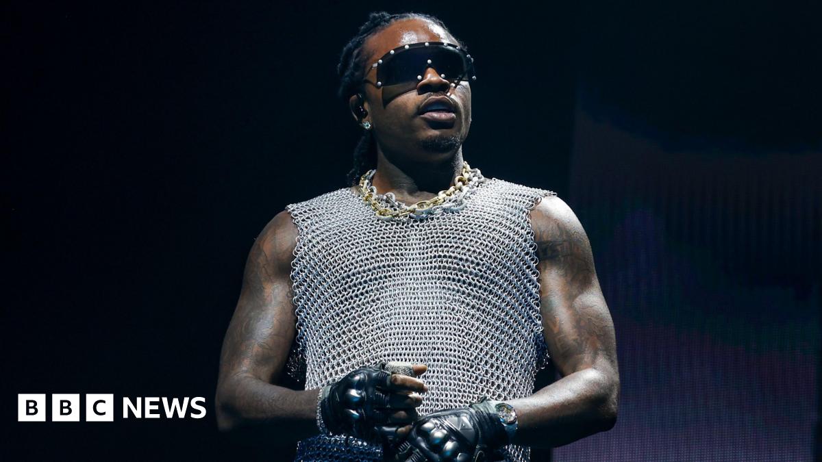 Gunna, dressed in gold and silver chains wears a silver vest top, black gloves and sunglasses while holding a mic