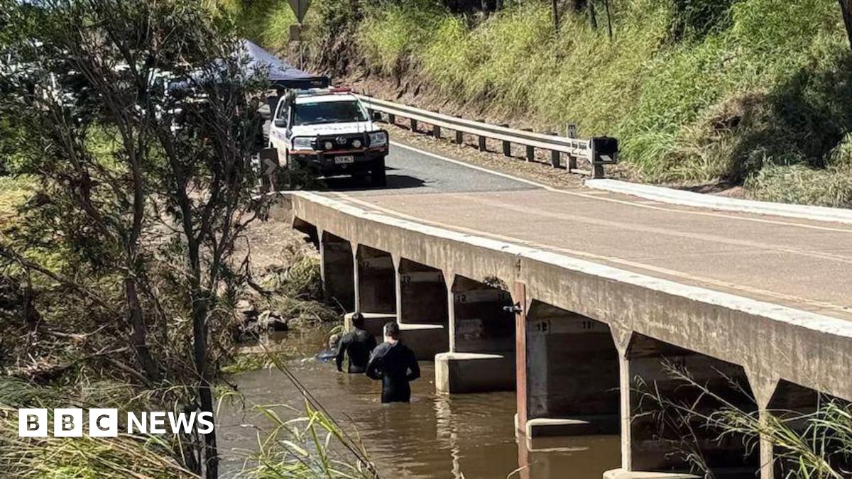 Two men in wet suits wade in a brown river running underneath a concrete road bridge with a rescue vehicle and tent on the road