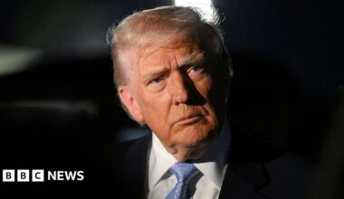 Trump wearing a black suit and blue tie looks into the distance. The background is black.