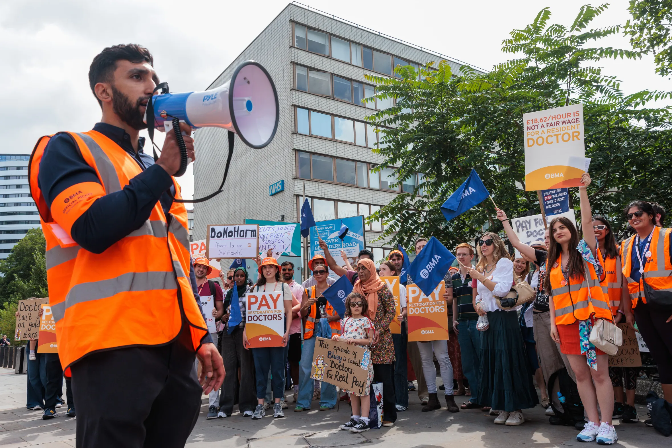 A BMA resident doctor speaks through a megaphone at a strike for better pay outside St Thomas' Hospital in London, surrounded by other picketing doctors holding signs and flags.