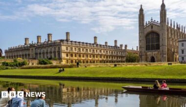 Three students sit in the foreground by the river Cam, opposite Kings College Cambridge on a sunny day. Two more students are in a punt on the river passing by in front of them.