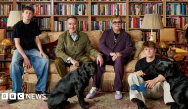Catherine Opie's photo of Elton and his family - Zachary, David, Elton and Elijah with the two pet black labradors in the foreground. They are seated on a sofa and rug in a library lined with bookshelves.