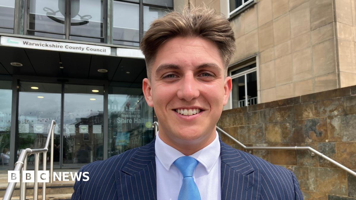 George Finch is looking directly at the camera smiling. He is wearing a navy pinstriped suit with a white shirt and a light blue tie. He is standing outside of the entrance to a building, in the background some steps and handrails can be seen, and behind them some automatic doors, above which is a sign that says Warwickshire County Council.
