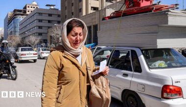A woman appears to be crying as she walks on a street in Tehran. She has a light coloured headscarf and a tan coat on. She is holding a piece of paper in her hand and has a rucksack on her shoulder. A motorcyclist is behind her and cars line the street to her left.