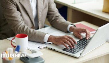 A close up of a business person in a brown suit and white shirt (we only see from the neck down) typing on a grey laptop on a desk. The desk has a white and red mug, a hole puncher and stationary.