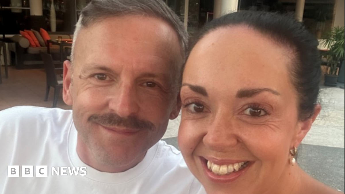 Laura and Paul Webster, a man with shirt grey hair and a mustache is wearing a white tshirt and looking at the camera with a slight smile, a woman next to him has dark brown hair pulled back and pearl earrings and looking at the camera with a wide smile