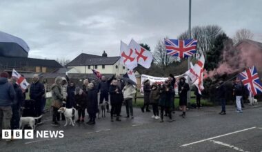 Protesters with St George's flags and Union Jacks, banners and flares gather in coats with dogs on the road outside the house.
