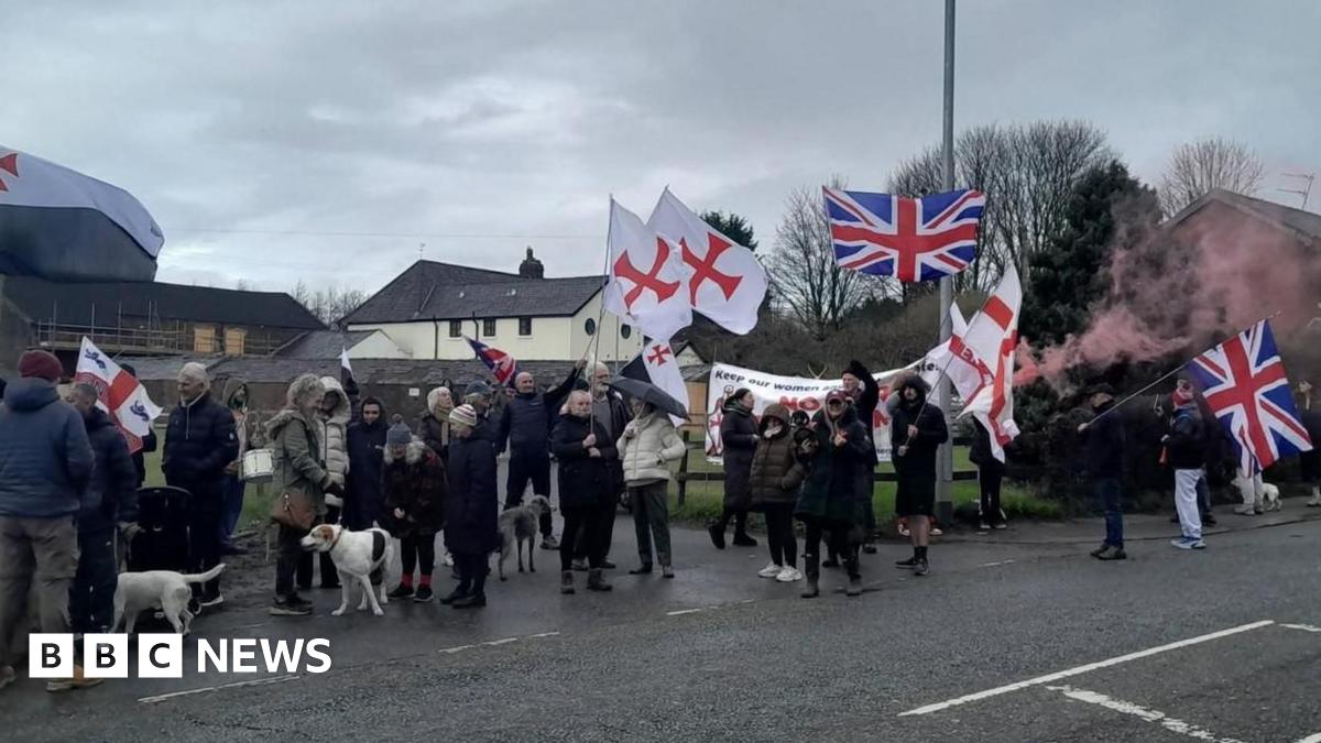 Protesters with St George's flags and Union Jacks, banners and flares gather in coats with dogs on the road outside the house.
