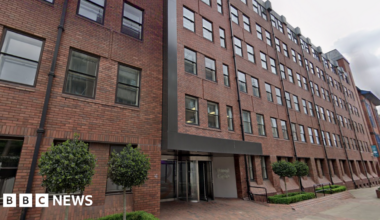 A five storey red brick office building with four small trees and small hedges outside. A glass revolving door entrance.