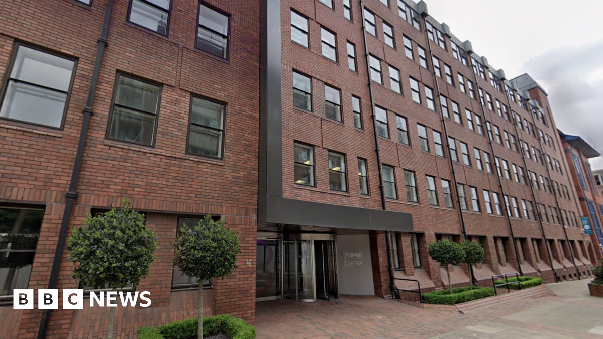 A five storey red brick office building with four small trees and small hedges outside. A glass revolving door entrance.