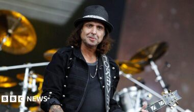 Phil looking at the camera playing guitar on stage. He wears a black hat, black t-shirt and a black jacket.