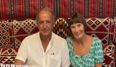 A man and a woman sat together on a sofa looking towards the camera.