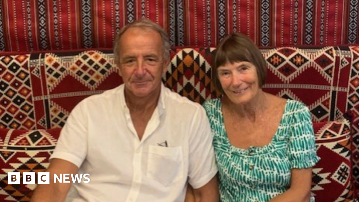 A man and a woman sat together on a sofa looking towards the camera.
