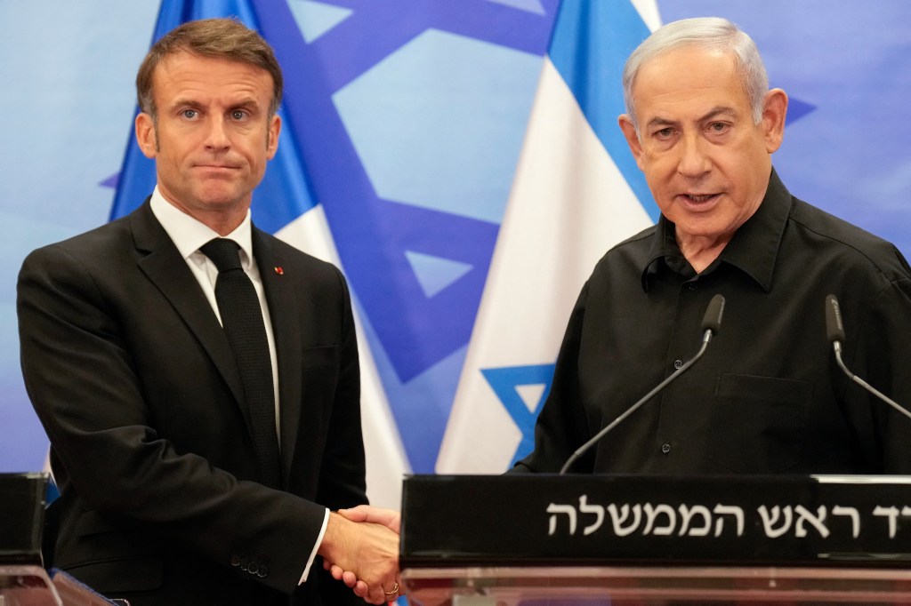 Israeli Prime Minister Benjamin Netanyahu (right) shakes hands with French President Emmanuel Macron (left) in front of an Israeli flag.